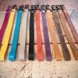 Op de afbeelding: Een verzameling gekleurde leren polsbandjes met zilverkleurige draaisluitingen. De bandjes zijn in verschillende kleuren, waaronder paars, geel, turquoise, grijs, zwart, koraal en bruin. Ze liggen op een houten ondergrond.