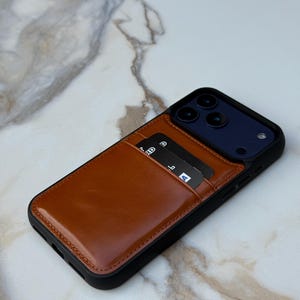 Peut inclure: Une coque de smartphone noire dot&eacute;e d'une pochette porte-cartes en cuir marron clair &agrave; l'arri&egrave;re. La pochette contient deux cartes bancaires noires avec du texte et des ic&ocirc;nes blancs visibles. La coque pr&eacute;sente une d&eacute;coupe pour le triple objectif.