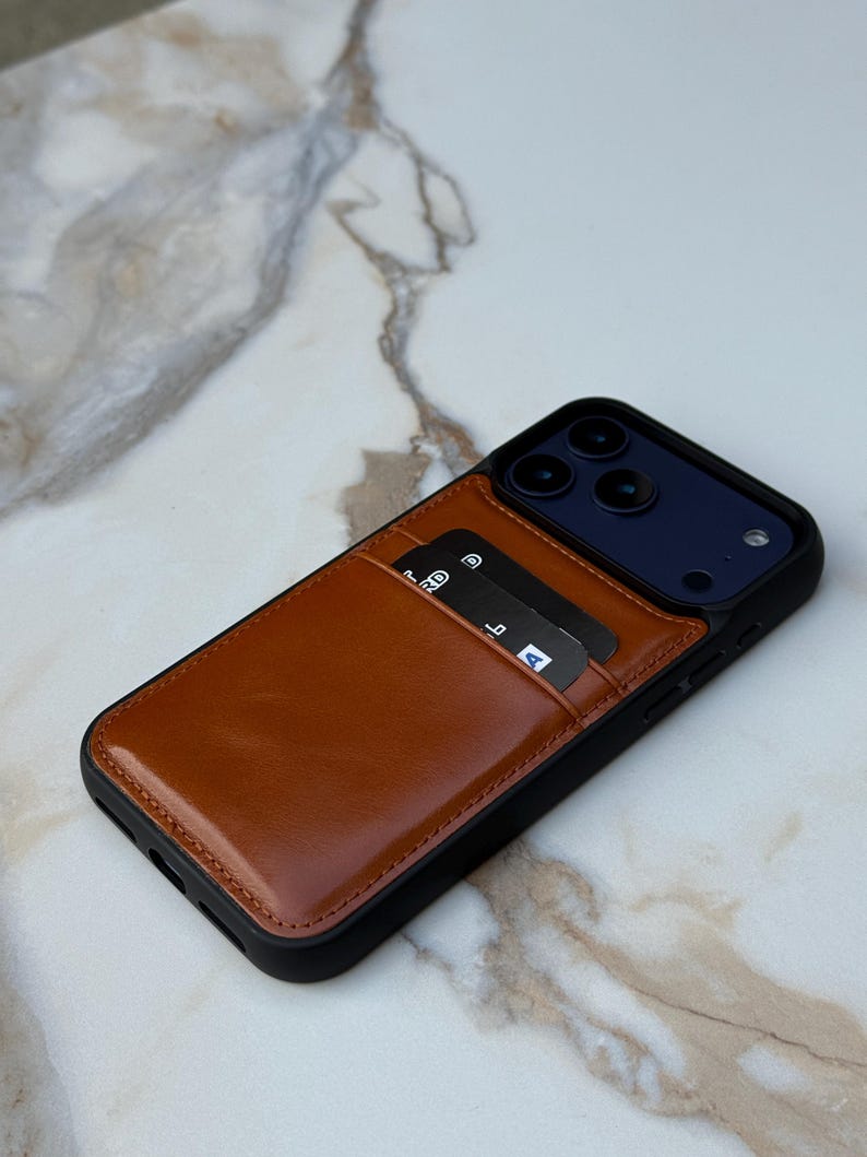 Peut inclure: Une coque de smartphone noire dot&eacute;e d'une pochette porte-cartes en cuir marron clair &agrave; l'arri&egrave;re. La pochette contient deux cartes bancaires noires avec du texte et des ic&ocirc;nes blancs visibles. La coque pr&eacute;sente une d&eacute;coupe pour le triple objectif.