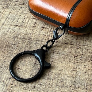 May include: A tan leather protective case for wireless earbuds featuring a black trim and a black metal lobster clasp keychain attached to the side. The case has a smooth texture and a secure closure mechanism with a circular ring for attachment.