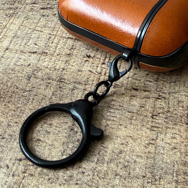 May include: A tan leather protective case for wireless earbuds featuring a black trim and a black metal lobster clasp keychain attached to the side. The case has a smooth texture and a secure closure mechanism with a circular ring for attachment.