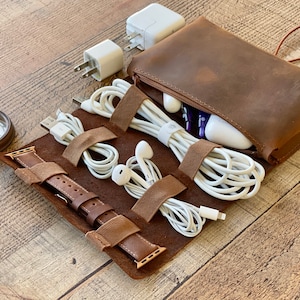 Puede incluir: Un organizador de viaje de cuero marrón con un bolsillo con cremallera y una sección enrollable con correas. Contiene cables de carga blancos, auriculares, una correa de reloj y una brújula. El organizador está sobre una superficie de madera.
