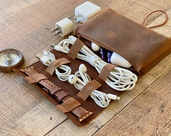 May include: A brown leather travel organizer with a zippered pouch and a roll-up section with straps. It holds white charging cables, earbuds, a watch band, and a compass. The organizer is on a wooden surface.