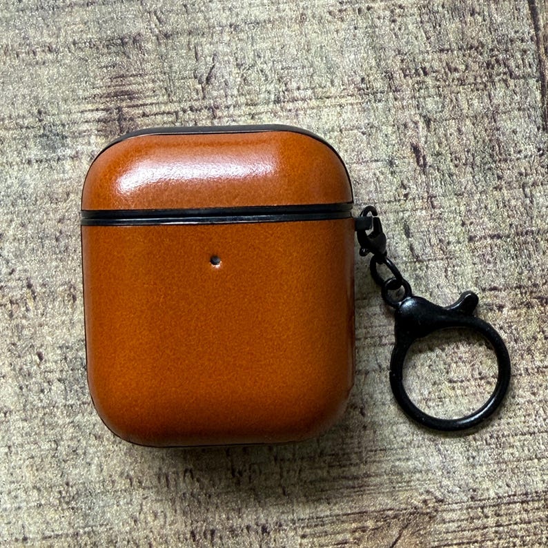 May include: A cognac-colored leather protective case for wireless earbuds featuring a black trim and a matching black metal keychain attachment with a circular ring. The case has a small circular cutout on the front for a status light indicator.
