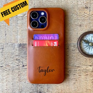 May include: A tan leather phone case with card slots, holding two credit cards. The case is personalised with the name "Taylor". The text "FREE CUSTOM" is in a yellow banner in the upper left corner.
