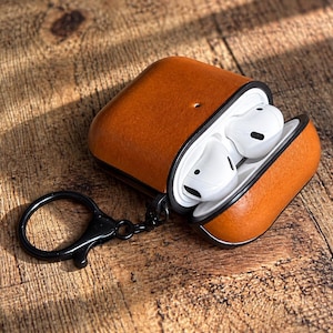 Puede incluir: Una funda protectora de cuero color canela para auriculares inalámbricos, con un forro interior negro y una anilla de metal negro para llaves unida al lateral. La funda está abierta, revelando dos auriculares blancos en el compartimento.