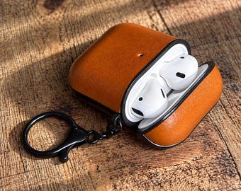 May include: A tan leather protective case for wireless earbuds, featuring a black interior lining and a black metal keychain ring attached to the side. The case is open, revealing two white wireless earbuds nestled inside the charging compartment.