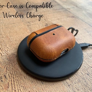 May include: A tan leather protective case for wireless earbuds resting on a black circular wireless charging pad. The text "Leather Case is Compatible With Wireless Charge" is visible in the upper left corner of the image.
