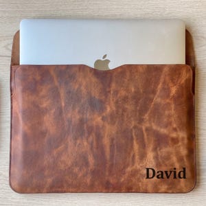 May include: A brown leather laptop sleeve, embossed with the name "David". The sleeve holds a silver laptop, with the Apple logo visible. The leather has a distressed, textured finish, giving it a vintage look.