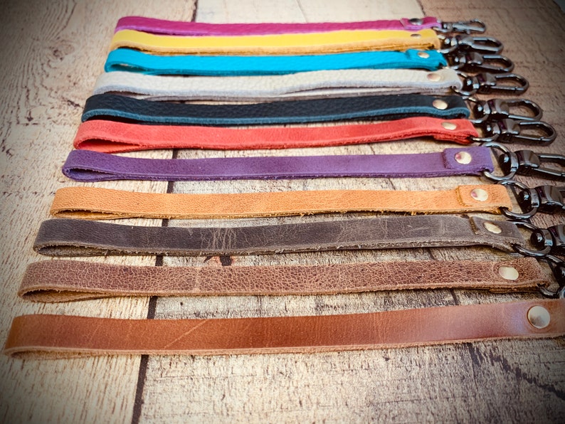 Op de afbeelding: Een verzameling gekleurde leren polsbandjes met metalen draaisluitingen. De bandjes zijn er in verschillende kleuren, waaronder roze, geel, turquoise, grijs, zwart, rood, paars, bruin, bruin en lichtbruin. Elke band heeft een andere kleur.