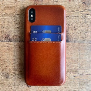 Peut inclure: Une coque de t&eacute;l&eacute;phone en cuir marron lisse dot&eacute;e d'une fente verticale contenant deux cartes bancaires bleues. La coque pr&eacute;sente une d&eacute;coupe pour un syst&egrave;me de double appareil photo en haut &agrave; gauche et des coutures visibles sur la poche.