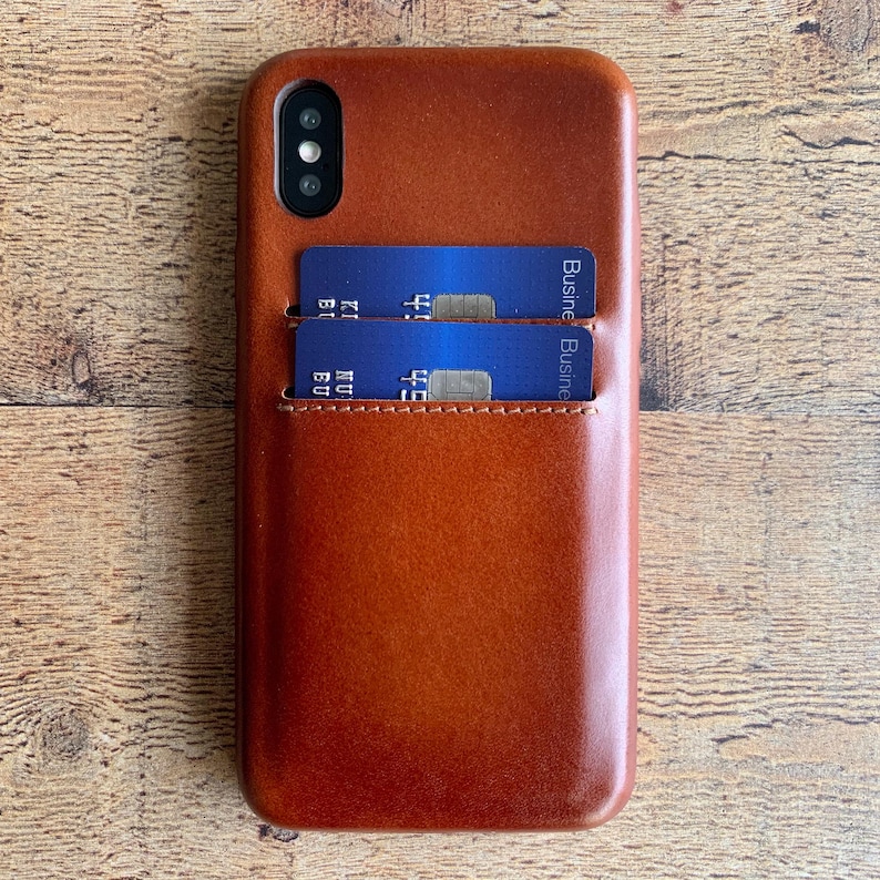 Peut inclure: Une coque de t&eacute;l&eacute;phone en cuir marron lisse dot&eacute;e d'une fente verticale contenant deux cartes bancaires bleues. La coque pr&eacute;sente une d&eacute;coupe pour un syst&egrave;me de double appareil photo en haut &agrave; gauche et des coutures visibles sur la poche.
