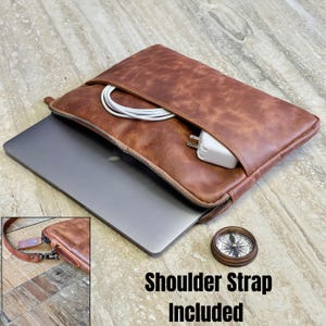 May include: A brown leather laptop sleeve with a zip, containing a laptop, charger, and cable. The text "Shoulder Strap Included" is visible. A small compass is next to the sleeve.