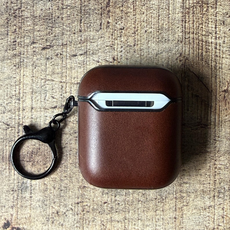 May include: A dark brown protective case for wireless earbuds featuring a smooth, matte finish and a white interior rim. The case includes a black metal circular keychain attachment connected to the side, providing a secure way to carry the device.