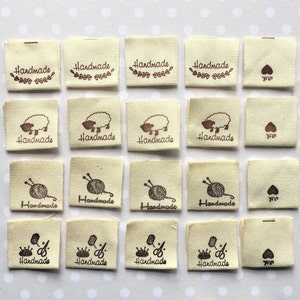 K&ouml;nnte beinhalten: Vierundzwanzig beigefarbene Stoff-Etiketten mit braunen Druckdesigns. Die Designs beinhalten ein Herz, ein Schaf, eine Wollkn&auml;uel mit Stricknadeln und eine Krone mit einer Schere. Die Etiketten sagen "Handmade".