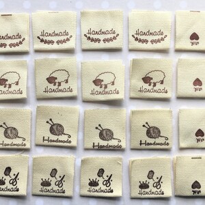 K&ouml;nnte beinhalten: Vierundzwanzig beigefarbene Stoff-Etiketten mit braunen Druckdesigns. Die Designs beinhalten ein Schaf, eine Wollkn&auml;uel mit Stricknadeln, eine Krone mit einer Schere und ein Herz. Die Etiketten sagen "Handmade".