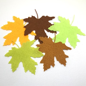 May include: A selection of felt maple leaves in shades of brown, green, and yellow. The leaves are arranged on a white surface, showcasing their detailed cut-out shapes and delicate stems. Suitable for autumn-themed crafts and decorations.