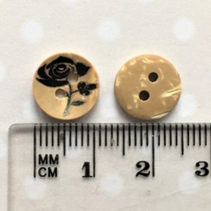 Puede incluir: Dos botones de madera, uno con un dise&ntilde;o de rosa negra y el otro con un patr&oacute;n de m&aacute;rmol beige. Ambos botones tienen un di&aacute;metro de aproximadamente 1 cm.