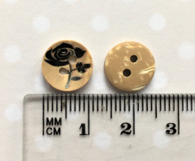 Puede incluir: Dos botones de madera, uno con un dise&ntilde;o de rosa negra y el otro con un patr&oacute;n de m&aacute;rmol beige. Ambos botones tienen un di&aacute;metro de aproximadamente 1 cm.