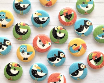 以下が含まれることがあります： 鳥のイラストが描かれた、カラフルな丸いボタンのコレクション。青、緑、黄色、赤の色合いで、白と黒のアクセントが施された様々な鳥のデザインが特徴です。各ボタンには縫い付け用の穴が2つあります。