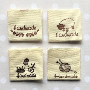 K&ouml;nnte beinhalten: Vier beigefarbene Stoff-Etiketten mit braunem Text und Bildern. Die Etiketten lauten "Handmade" mit verschiedenen Bildern, darunter ein Schaf, eine Krone, eine Schere und eine Wollkn&auml;uel mit Nadeln.