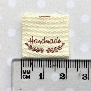 K&ouml;nnte beinhalten: Ein wei&szlig;es Stofflabel mit brauner Stickerei, auf dem "Handmade" steht, darunter ein Blattdesign. Das Label wird an einem Lineal gemessen und ist etwa 3,8 cm breit.