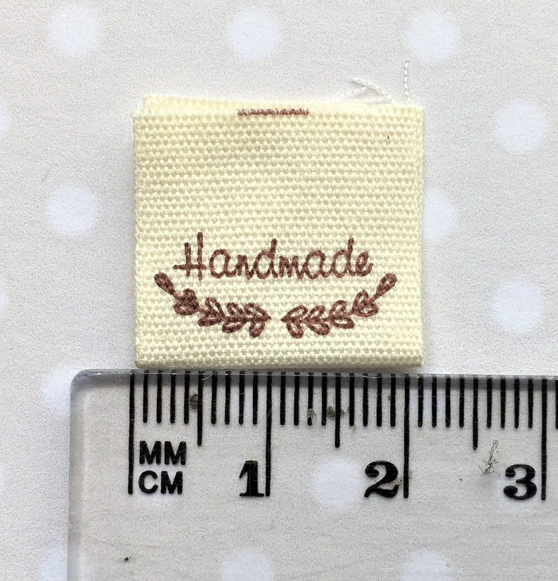 K&ouml;nnte beinhalten: Ein wei&szlig;es Stofflabel mit brauner Stickerei, auf dem "Handmade" steht, darunter ein Blattdesign. Das Label wird an einem Lineal gemessen und ist etwa 3,8 cm breit.