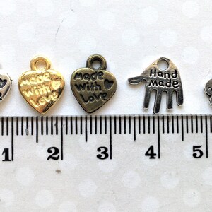 May include: A collection of small metal charms in various shapes and finishes. The charms include heart shapes with "Made with Love" text, a hand shape, and a flower shape, all with "Hand Made" text. A ruler with centimeter markings is in the background.