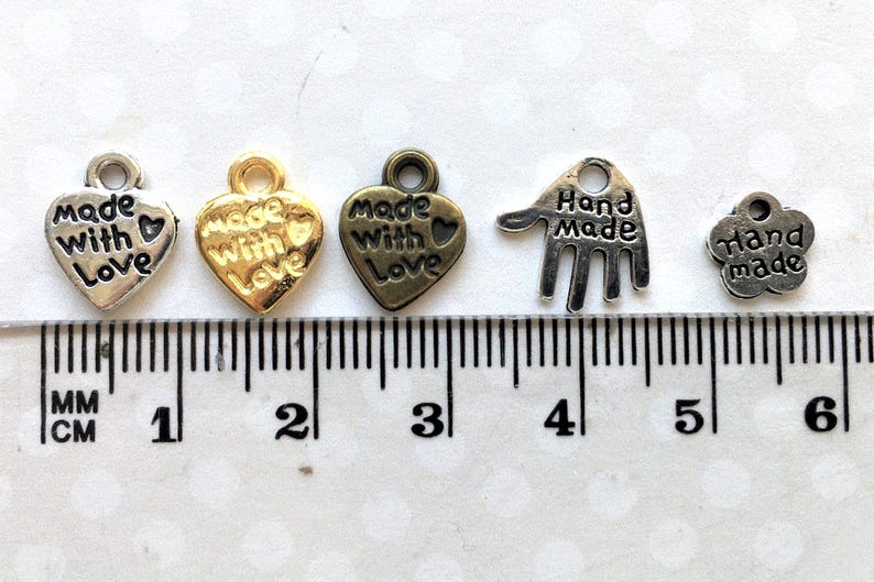 May include: A collection of small metal charms in various shapes and finishes. The charms include heart shapes with "Made with Love" text, a hand shape, and a flower shape, all with "Hand Made" text. A ruler with centimeter markings is in the background.