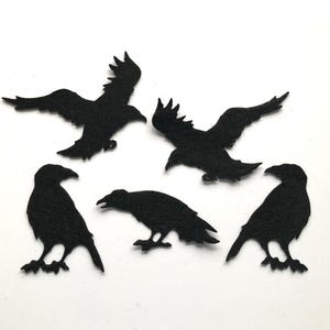 May include: Five black felt raven silhouettes in different poses, including flying and perched. The ravens are cut from a dark, textured material and set against a white background. These decorative cutouts are suitable for crafting or seasonal decor.