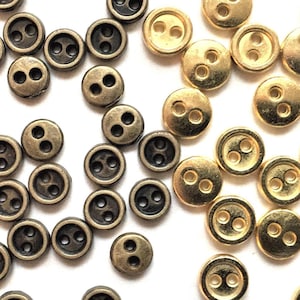 May include: A selection of small, round buttons in antique brass and dark bronze. Each button features two holes for sewing. The buttons are scattered on a white surface, highlighting their metallic sheen and simple design.