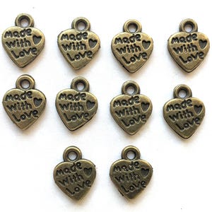 May include: A collection of bronze-colored heart-shaped charms. Each charm has the words "Made With Love" embossed on the front. The charms have a small loop at the top for attaching to jewelry or other crafts.