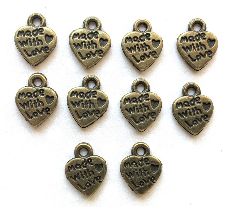 May include: A collection of bronze-colored heart-shaped charms. Each charm has the words "Made With Love" embossed on the front. The charms have a small loop at the top for attaching to jewelry or other crafts.