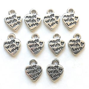 May include: A collection of silver-colored heart-shaped charms. Each charm has the words "Made With Love" engraved on the front. The charms have a small loop at the top for attaching to jewelry or other crafts.