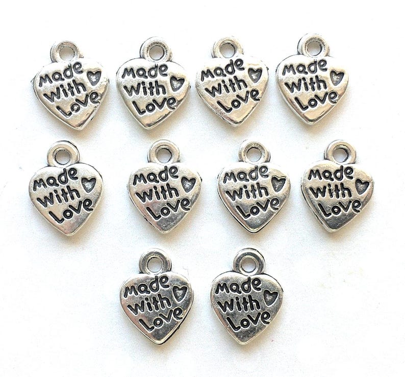 May include: A collection of silver-colored heart-shaped charms. Each charm has the words "Made With Love" engraved on the front. The charms have a small loop at the top for attaching to jewelry or other crafts.