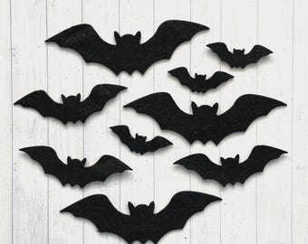 以下が含まれることがあります： さまざまなサイズの8つの黒いコウモリ型の切り抜き。各コウモリは、尖った耳と広げた翼を持つ対称的なシルエットが特徴で、白い木目調の背景に散らばったパターンで配置されています。