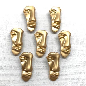 May include: Gold-coloured face-shaped embellishments with a matte finish. The decorative pieces feature a stylized human face with defined features. These small, detailed faces are ideal for crafting and jewellery making.