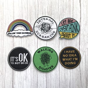 May include: A selection of embroidered patches featuring humorous phrases and designs. Includes a rainbow with "Follow Your Daydream", a black patch with "It's OK to Not Be OK", and a green patch with "Walking Disaster". Other patches include "Internal Screaming", "Let Me Overthink About It", and "I Have No Idea What I'm Doing".