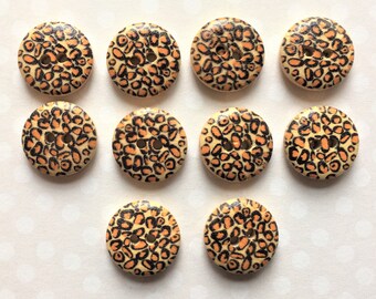 May include: A collection of ten round wooden buttons with a leopard print design. The buttons have a light beige background with orange and black spots. Each button has two holes for sewing.
