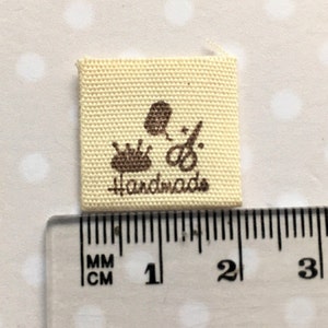 K&ouml;nnte beinhalten: Ein beigefarbenes Stofflabel mit braunen N&auml;hten und dem aufgedruckten Wort "handmade". Das Label zeigt au&szlig;erdem drei braune Symbole: eine Garnrolle, eine Schere und ein Blatt.