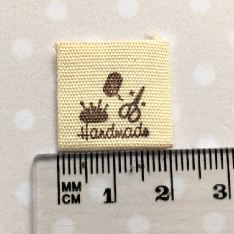 K&ouml;nnte beinhalten: Ein beigefarbenes Stofflabel mit braunen N&auml;hten und dem aufgedruckten Wort "handmade". Das Label zeigt au&szlig;erdem drei braune Symbole: eine Garnrolle, eine Schere und ein Blatt.