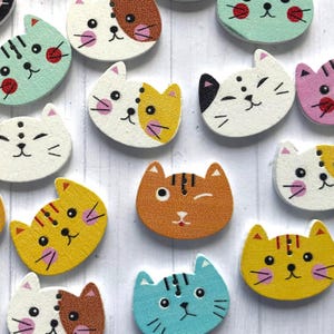 May include: Assortment of wooden cat face buttons in various colours, including white, blue, yellow, orange, and pink. The buttons feature different cat expressions, such as winking, smiling, and frowning.