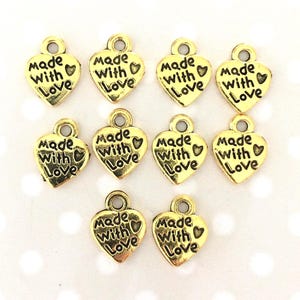 May include: Gold-colored heart-shaped charms with the words "Made With Love" inscribed. Each charm has a small loop at the top for attaching to jewelry or crafts. The charms are arranged on a light background.