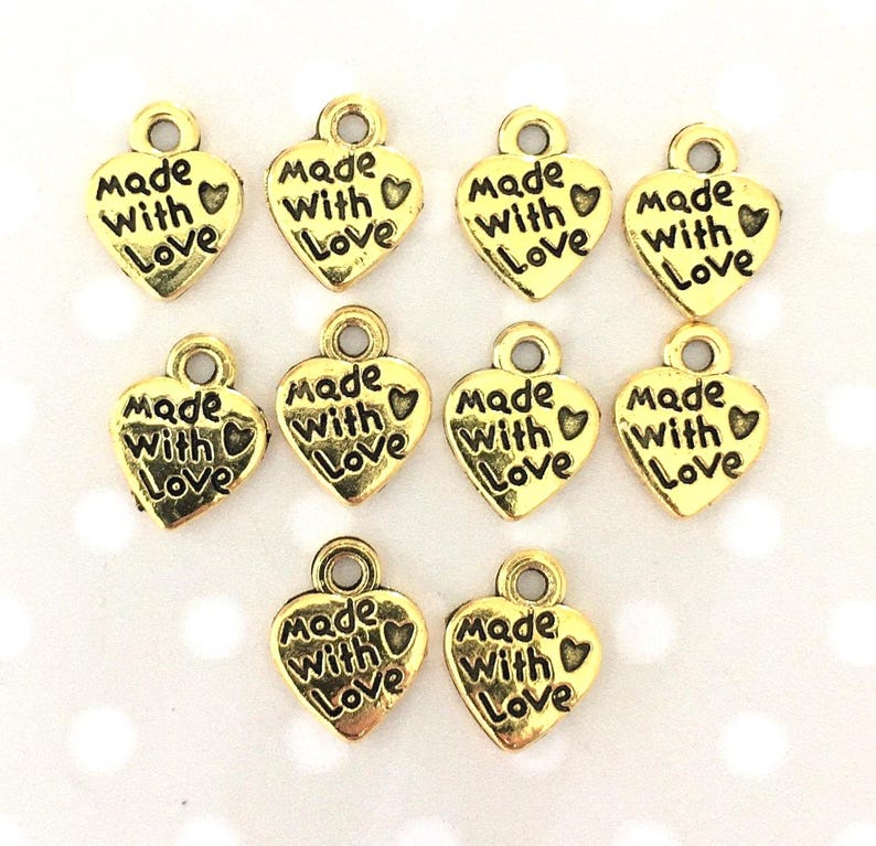 May include: Gold-colored heart-shaped charms with the words "Made With Love" inscribed. Each charm has a small loop at the top for attaching to jewelry or crafts. The charms are arranged on a light background.