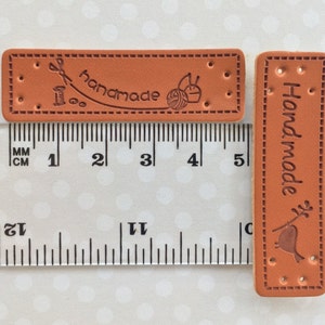 May include: Two brown leather labels with the word "Handmade" embossed on them. One label has a sewing needle, thread, and yarn ball design. Both labels have stitching around the edges and four holes for attaching.