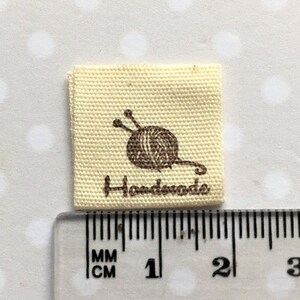 K&ouml;nnte beinhalten: Ein beigefarbenes Stofflabel mit einer braunen Wollkn&auml;uel- und Stricknadelgrafik und dem aufgedruckten Wort "Handmade". Das Etikett ist 2,5 cm quadratisch.