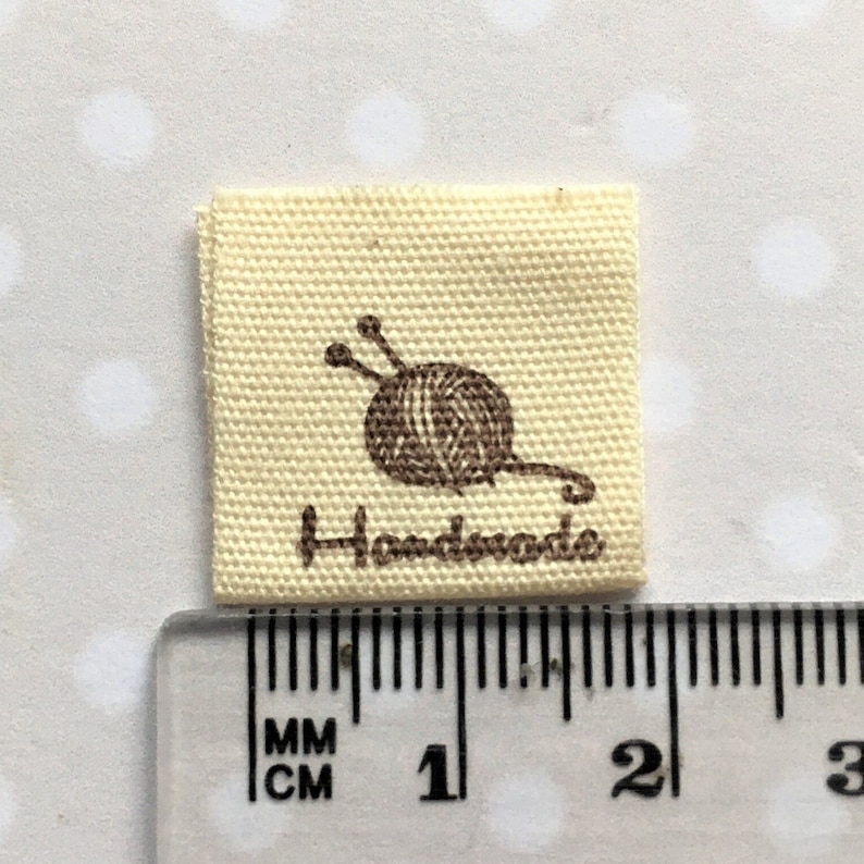 K&ouml;nnte beinhalten: Ein beigefarbenes Stofflabel mit einer braunen Wollkn&auml;uel- und Stricknadelgrafik und dem aufgedruckten Wort "Handmade". Das Etikett ist 2,5 cm quadratisch.