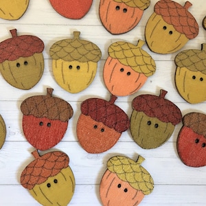 May include: A collection of 15 wooden acorn buttons in various shades of brown, orange, and yellow. The buttons have a raised acorn cap and two holes for sewing.