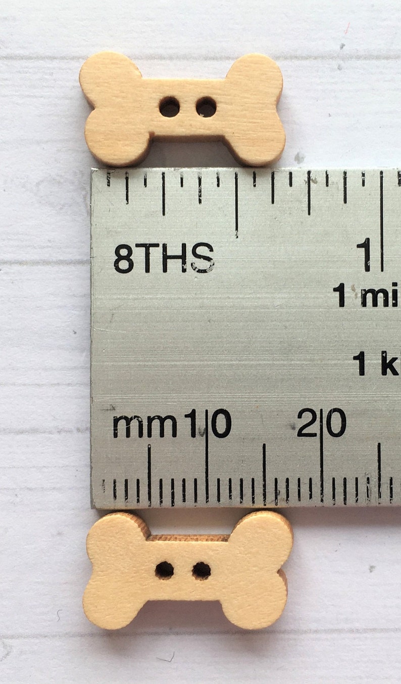 May include: Two wooden bone-shaped buttons with two holes each. The buttons are light brown and measure approximately 1 inch in length.