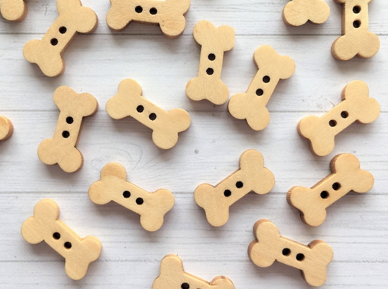 May include: A collection of wooden bone-shaped buttons with two holes each. The buttons are a light brown color and are arranged in a scattered pattern on a white wooden surface.